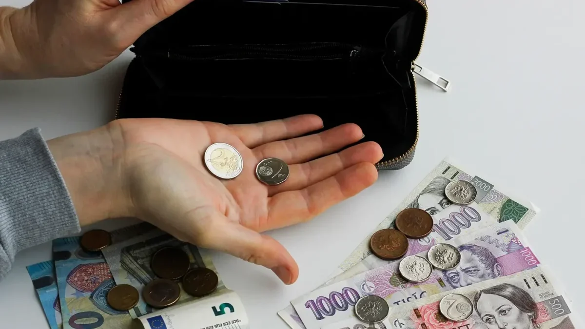 Člověk v ruce drží dvě mince a na stole leží další mince a papírové bankovky