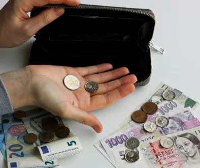 Člověk v ruce drží dvě mince a na stole leží další mince a papírové bankovky