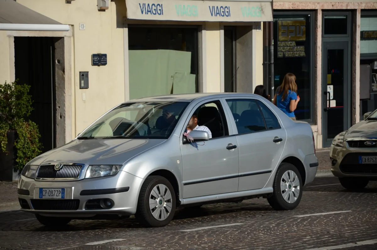 shutterstock_2239622229 1 | TVGURU.cz Silver car on a cobbled street in front of row of shops; a woman in blue walks by in the background.