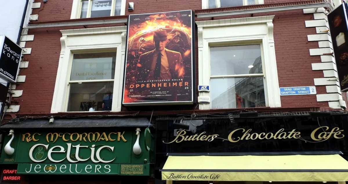 shutterstock_2332446645 1 | TVGURU.cz Brick storefront with a large Oppenheimer movie poster mounted between two windows of a red-brick building; signs for Celtic Jewellers and Butler's Chocolate Café below.