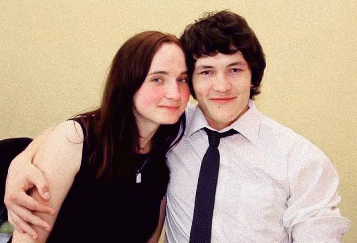 JB7649c7_kuc | TVGURU.cz Two young adults pose closely together, arm around each other, smiling at the camera against a beige wall; woman in a black top, man in a white shirt and dark tie.