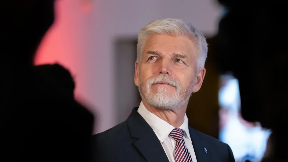 Senior man with gray hair and beard in a dark suit and red striped tie, looking to the right at a formal event.