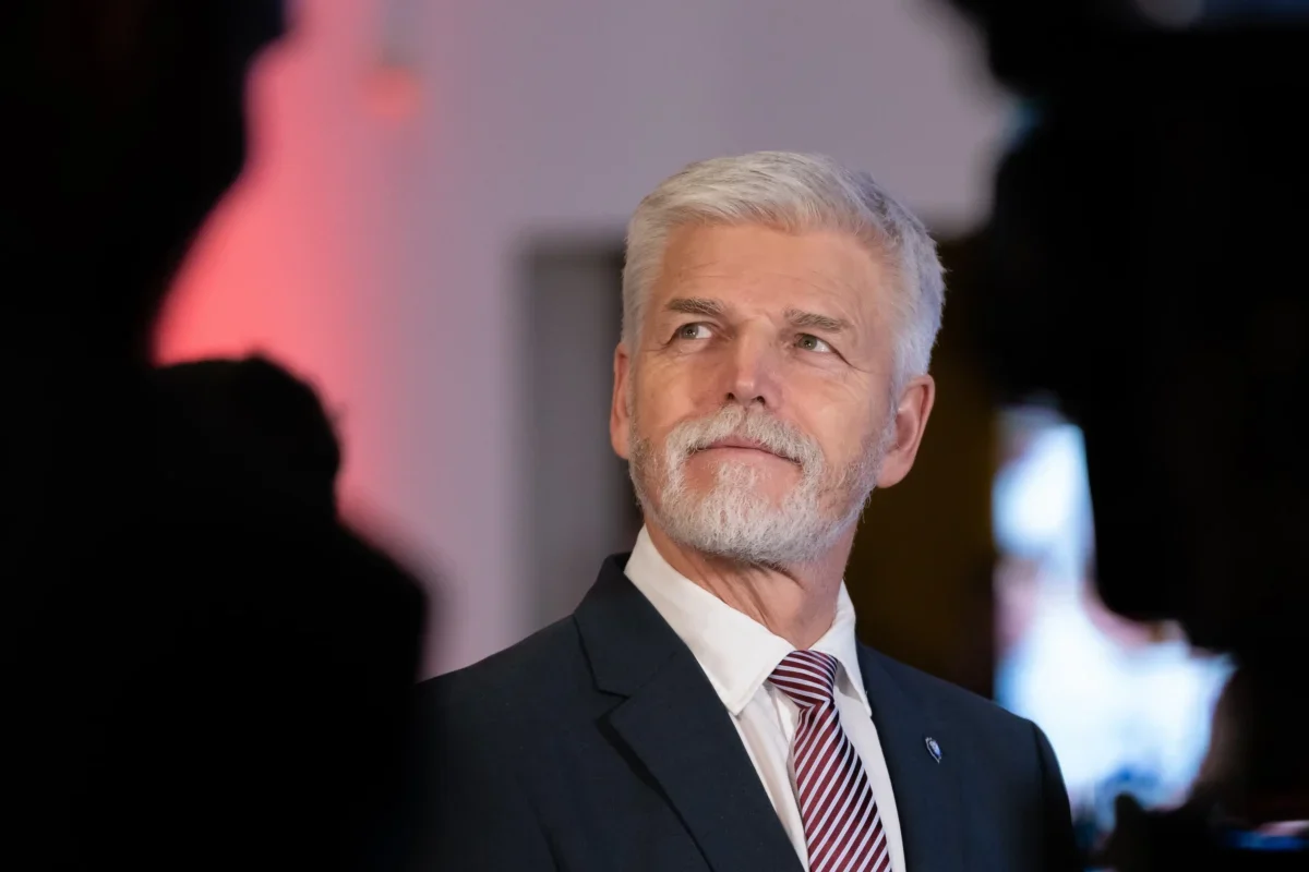 Senior man with gray hair and beard in a dark suit and red striped tie, looking to the right at a formal event.