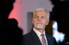Senior man with gray hair and beard in a dark suit and red striped tie, looking to the right at a formal event.