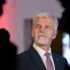 Senior man with gray hair and beard in a dark suit and red striped tie, looking to the right at a formal event.