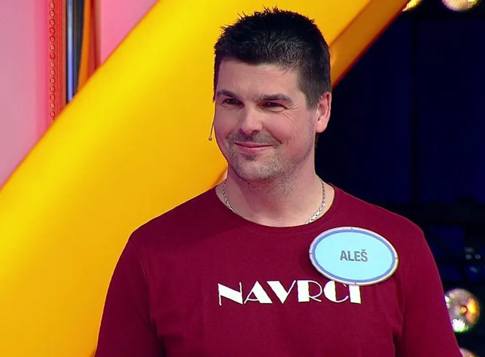 5c8bb787-a491-4c99-bb17-1334474fb7a1 | TVGURU.cz Smiling man with short dark hair, wearing a maroon shirt and a blue name badge that reads Aleš on a colorful stage.