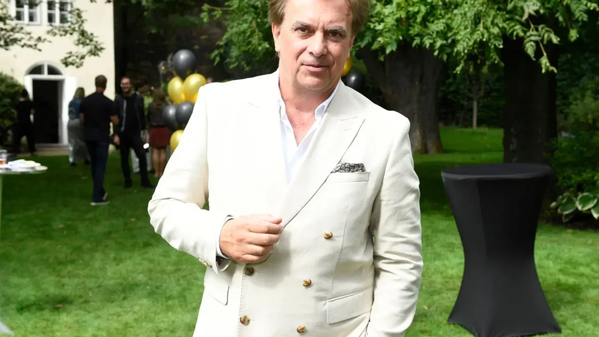 Man wearing a light beige blazer posing outdoors at a garden party, with balloons and guests in the background.