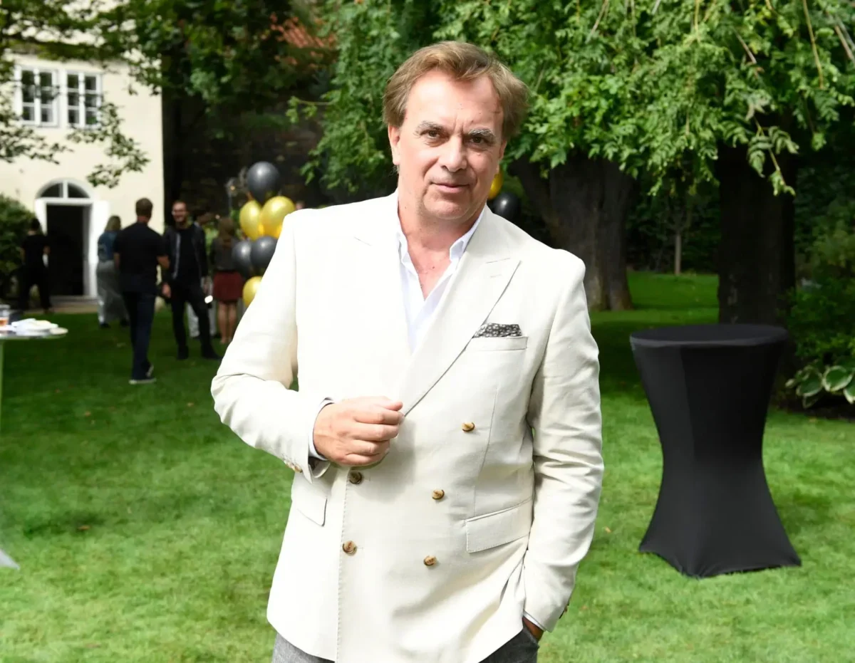 Man wearing a light beige blazer posing outdoors at a garden party, with balloons and guests in the background.