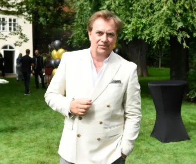 Man wearing a light beige blazer posing outdoors at a garden party, with balloons and guests in the background.