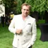 Man wearing a light beige blazer posing outdoors at a garden party, with balloons and guests in the background.