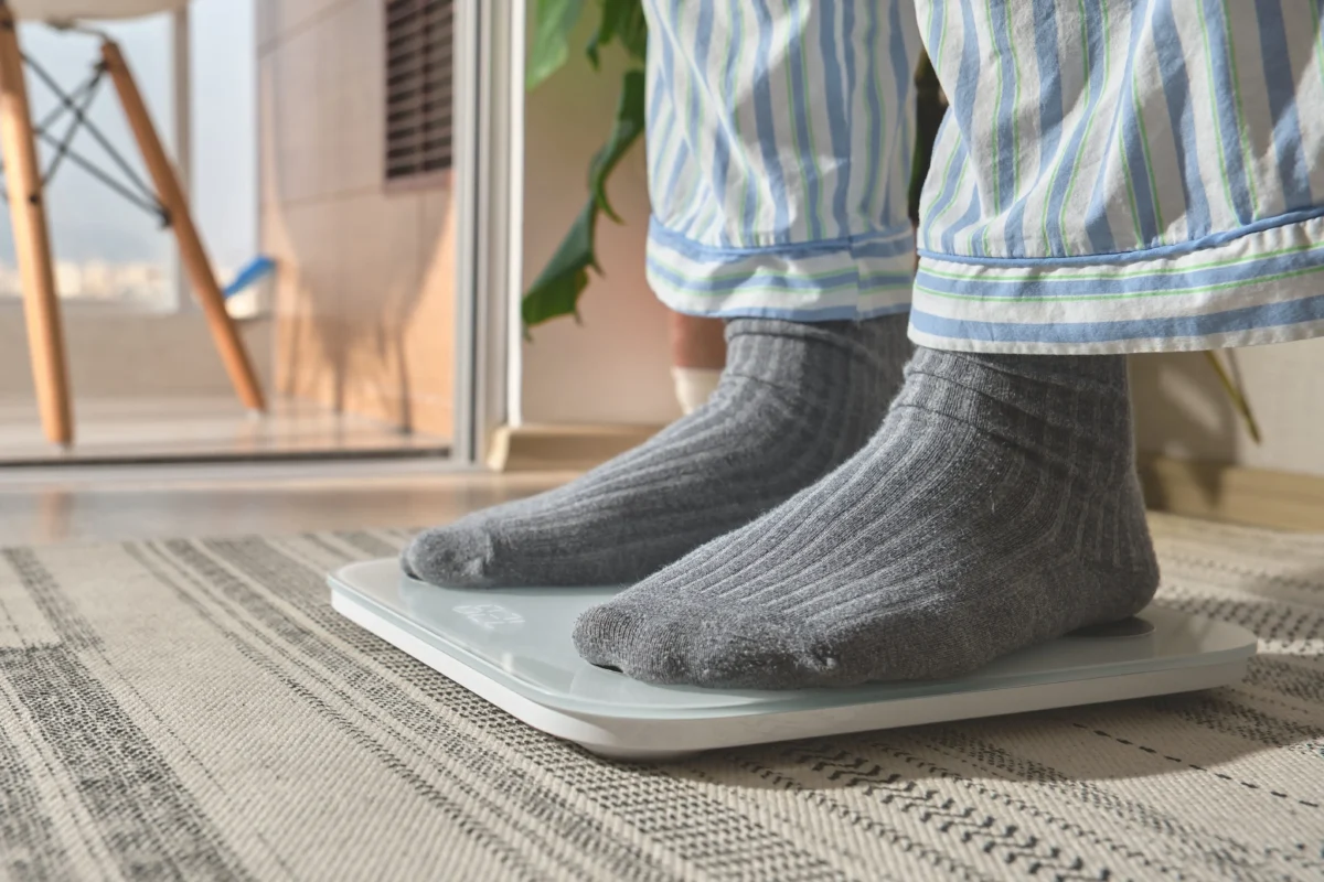 shutterstock_2715187069 1 | TVGURU.cz Person in striped clothing and gray socks stands on a glass‑top digital scale on a patterned rug inside a home, near a sliding door/window.