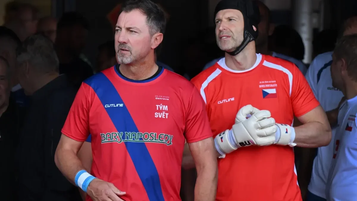 Two adult athletes in red jerseys with a blue diagonal sash, standing side by side, looking off to the left, wearing gloves.