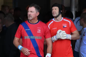 Two adult athletes in red jerseys with a blue diagonal sash, standing side by side, looking off to the left, wearing gloves.