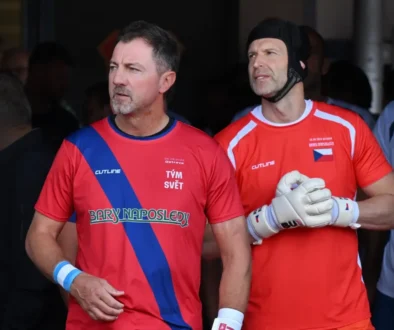 Two adult athletes in red jerseys with a blue diagonal sash, standing side by side, looking off to the left, wearing gloves.