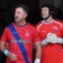 Two adult athletes in red jerseys with a blue diagonal sash, standing side by side, looking off to the left, wearing gloves.