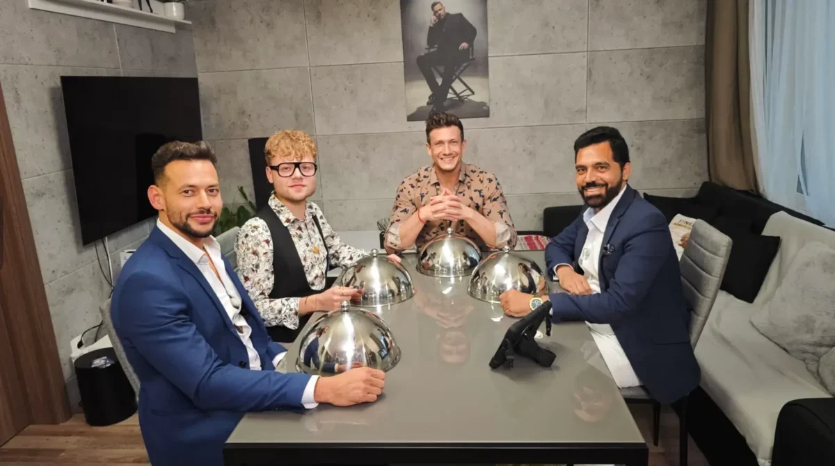 Four men in suits sit around a glass dining table with shiny silver cloche domes, posing for a photo in a modern room with a poster on the wall behind them.