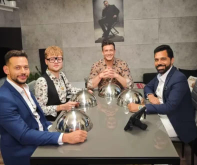 Four men in suits sit around a glass dining table with shiny silver cloche domes, posing for a photo in a modern room with a poster on the wall behind them.