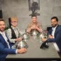 Four men in suits sit around a glass dining table with shiny silver cloche domes, posing for a photo in a modern room with a poster on the wall behind them.
