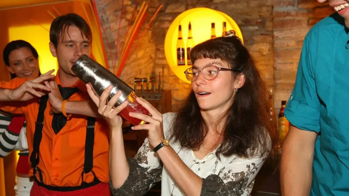 Žena s delšími tmavými kudrnatými vlasy a brýlemi drží v ruce šejkr a třese jím