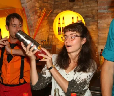 Žena s delšími tmavými kudrnatými vlasy a brýlemi drží v ruce šejkr a třese jím