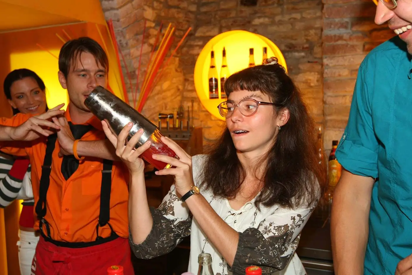 Žena s delšími tmavými kudrnatými vlasy a brýlemi drží v ruce šejkr a třese jím