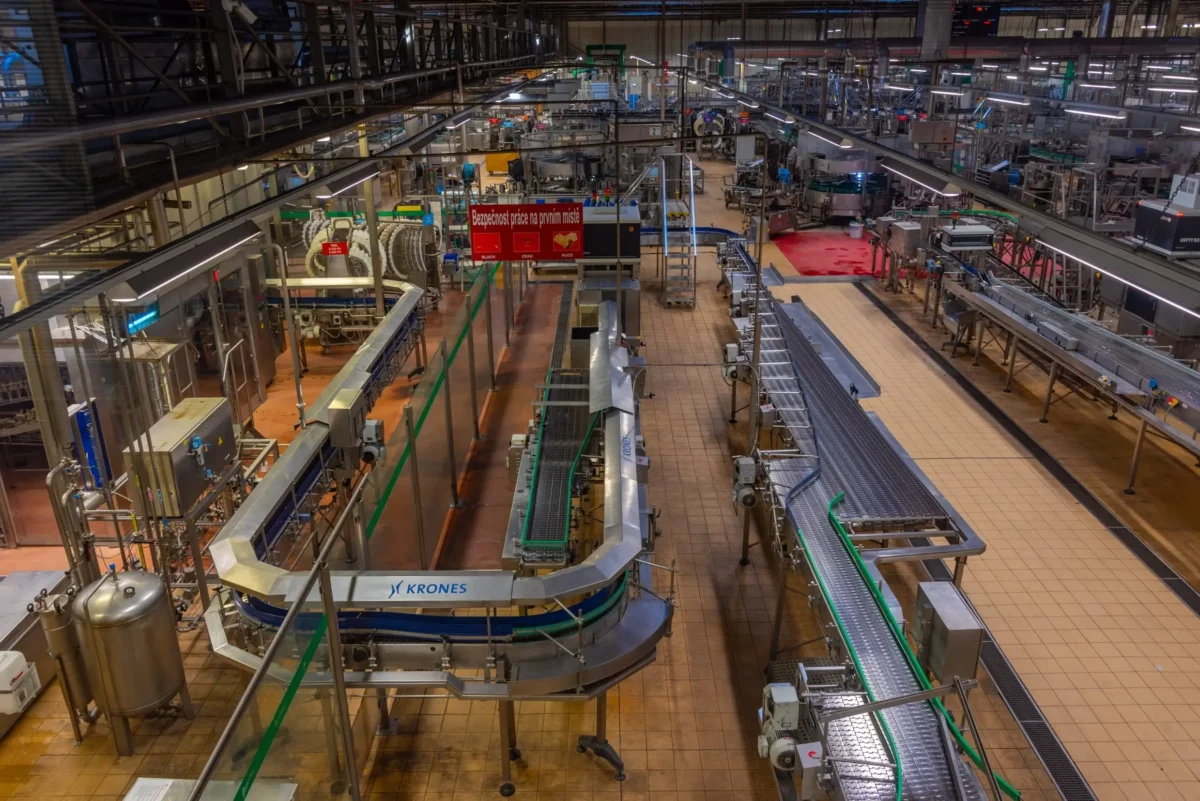 shutterstock_2674198511 1 | TVGURU.cz Wide view of a large beverage bottling plant with conveyor belts, stainless steel machinery, and overhead piping
