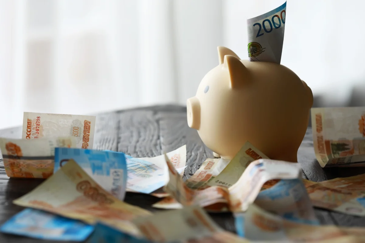 shutterstock_2794592729 1 | TVGURU.cz Beige piggy bank with a 2000-ruble bill inserted into its slot, surrounded by scattered banknotes on a table.