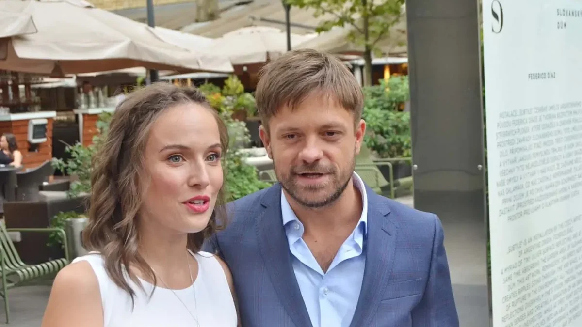 Smiling couple standing close together outdoors in a plaza, woman in a white sleeveless top and the man in a blue suit, with cafe umbrellas and greenery in the background.