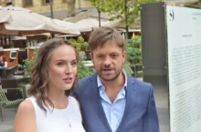 Smiling couple standing close together outdoors in a plaza, woman in a white sleeveless top and the man in a blue suit, with cafe umbrellas and greenery in the background.