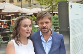 Smiling couple standing close together outdoors in a plaza, woman in a white sleeveless top and the man in a blue suit, with cafe umbrellas and greenery in the background.