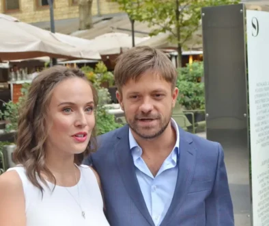 Smiling couple standing close together outdoors in a plaza, woman in a white sleeveless top and the man in a blue suit, with cafe umbrellas and greenery in the background.