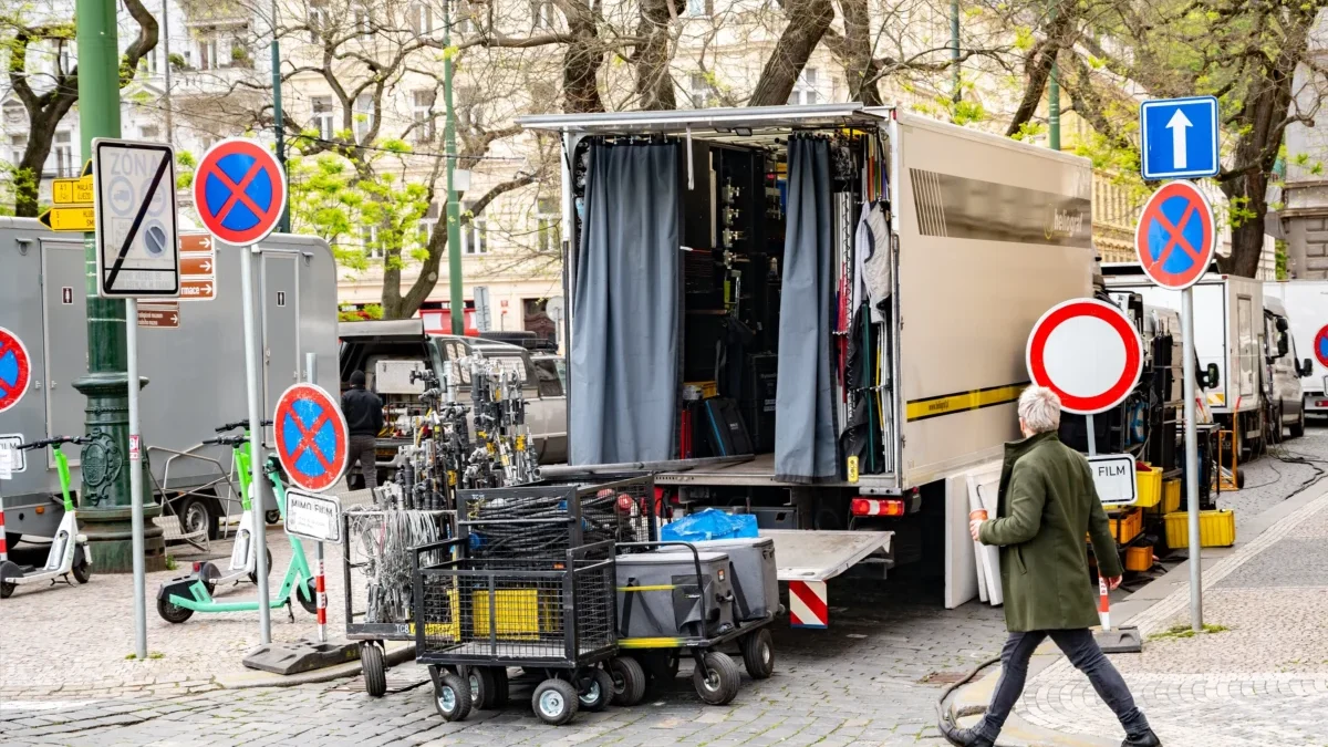 A street scene with a large beige film truck open for gear, surrounded by carts, cables and signs, and a man in a green coat walking by.