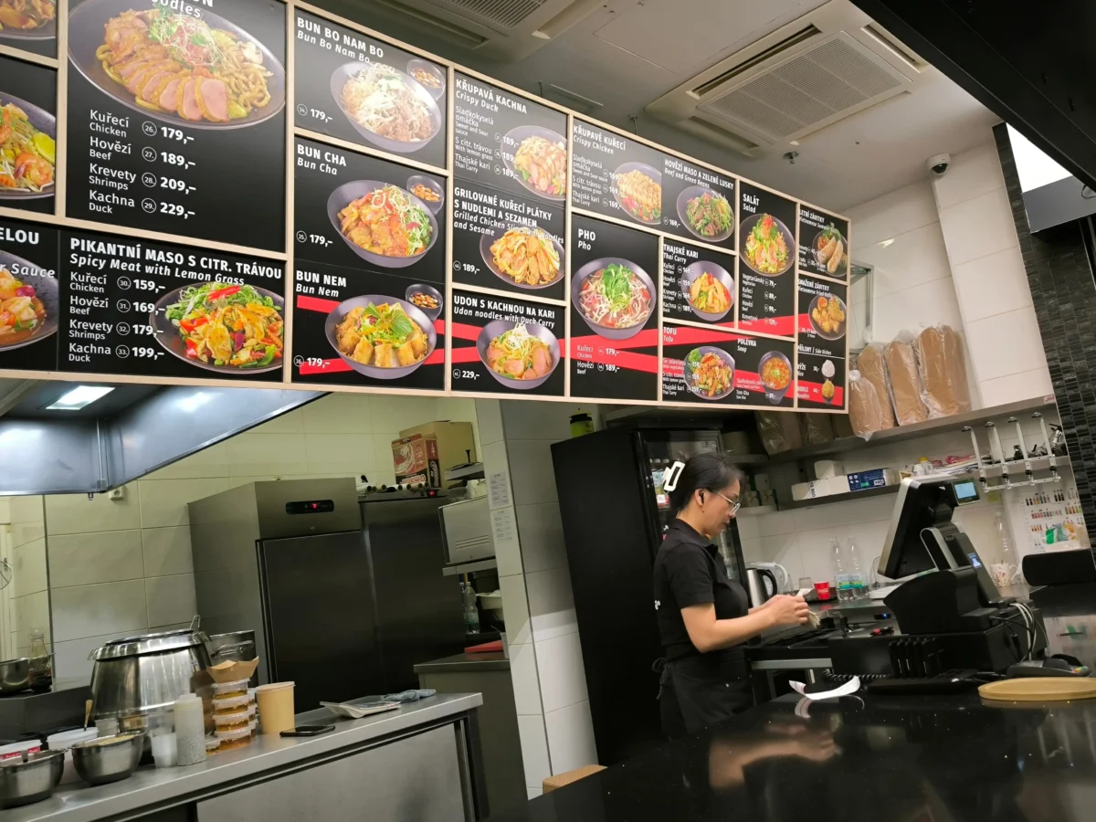 shutterstock_2784093919 1 | TVGURU.cz Wall of illuminated menu boards with photos and prices, above a stainless counter in a fast-casual restaurant; a staff member stands at the register.