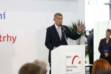 Muž stojí za bílým řečnickým pultíkem a gestikuluje