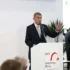 Muž stojí za bílým řečnickým pultíkem a gestikuluje