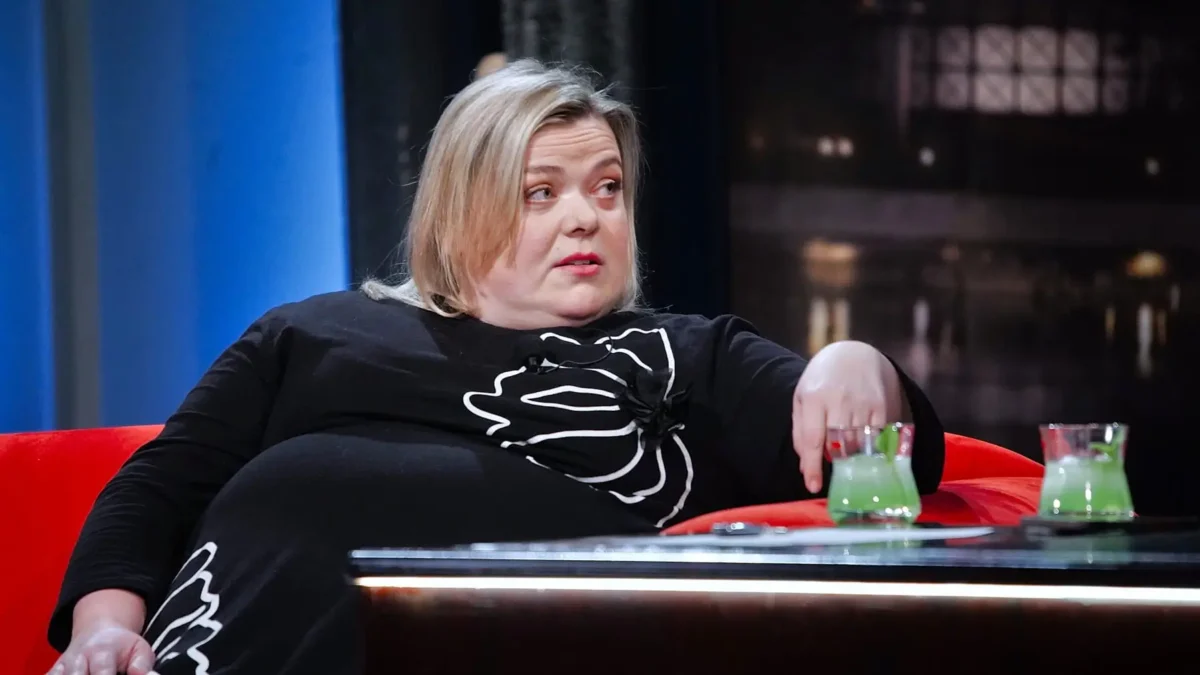 profimedia-1066152000 1 | TVGURU.cz Woman with blonde hair seated on a red chair, wearing a black dress with a white abstract pattern, resting her arm on a glass table with two green drinks.