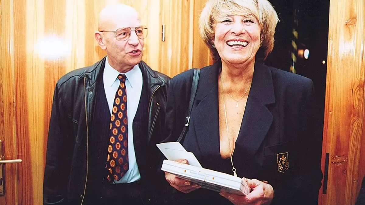 Žena v černém saku stojí v popředí před starším mužem a v ruce drží knihu