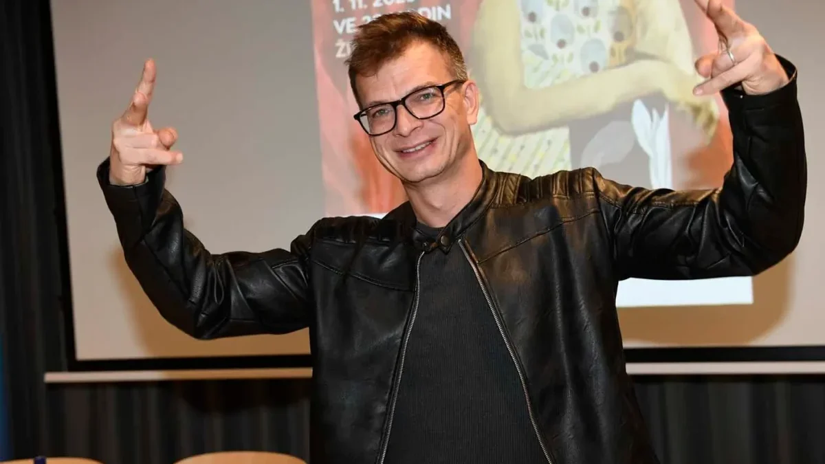 Muž s krátkými vlasy a v černém oblečení stojí před promítacím plátnem