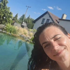 536618985_18072451154021580_2304149669123633073_n | TVGURU.cz Smiling woman with wet hair leaning into the frame beside a clear blue pool and a modern house in the background on a sunny day