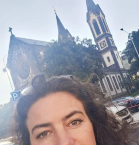 653709059_26382398894687717_5140841541139179520_n | TVGURU.cz Close-up of a person’s face outdoors with a church’s twin spires behind them.
