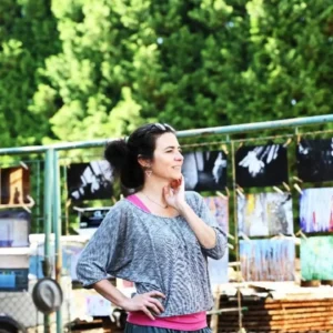 719828212_18102538040021580_4005336673981159621_n | TVGURU.cz Woman stands outdoors at an art market, hand on hip, smiling, with colorful paintings hanging on a fence in the background.