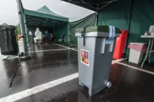 Medical testing tent with staff in PPE sitting at a table inside a green canopy structure in a parking lot.