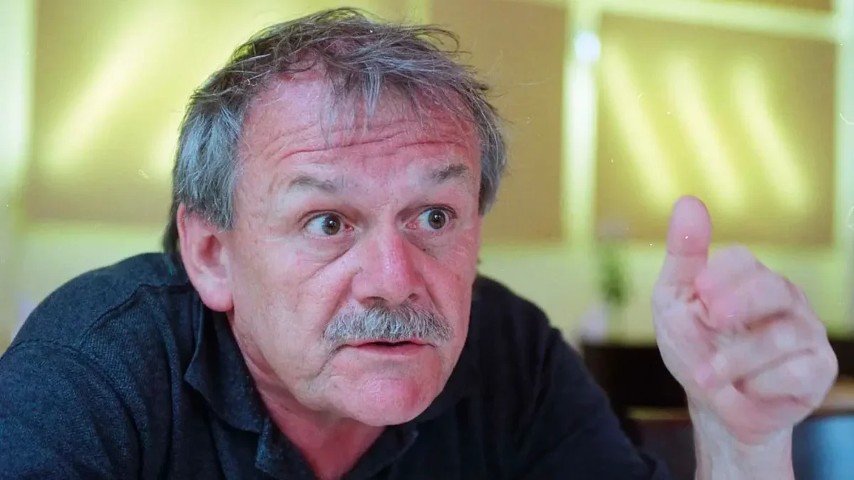 Starší muž s šedivými vlasy a šedivým knírkem na sobě má černé triko s límečkem a gestikuluje levou rukou