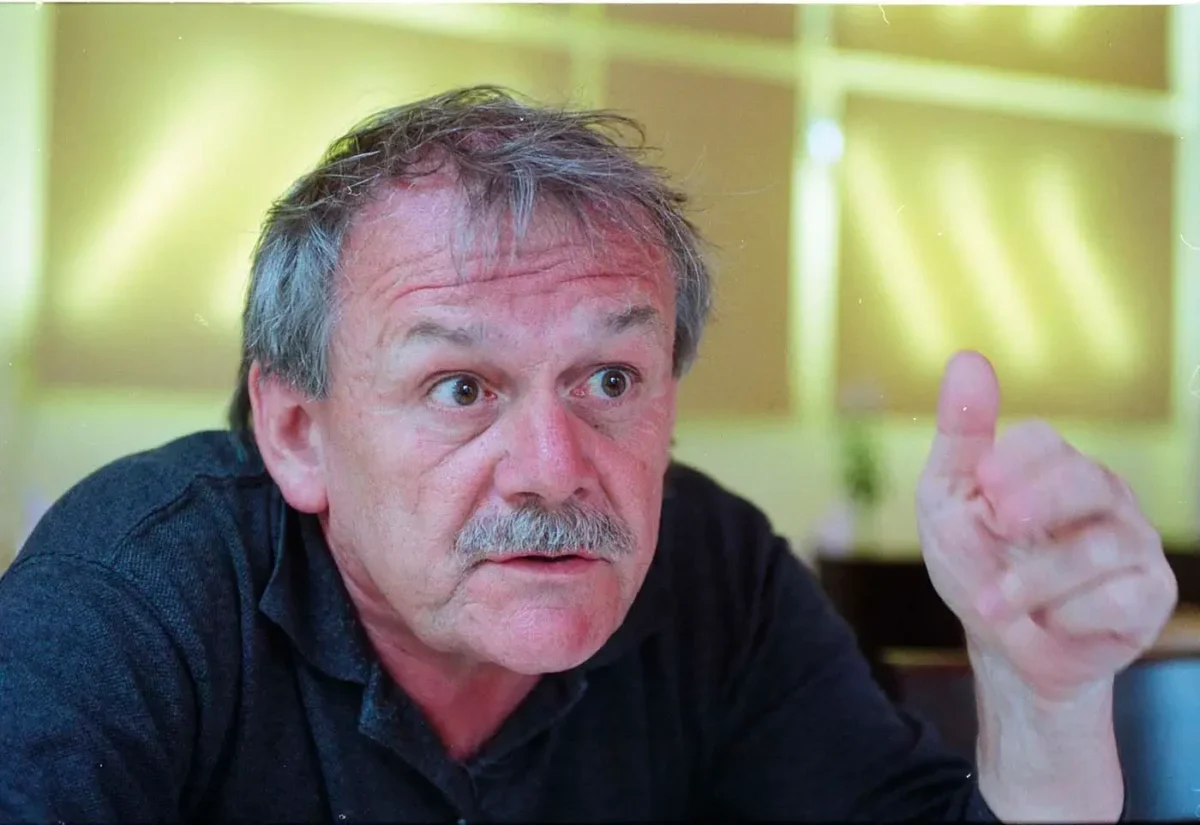 Starší muž s šedivými vlasy a šedivým knírkem na sobě má černé triko s límečkem a gestikuluje levou rukou