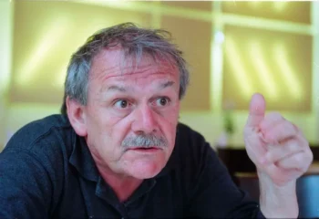 Starší muž s šedivými vlasy a šedivým knírkem na sobě má černé triko s límečkem a gestikuluje levou rukou