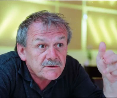 Starší muž s šedivými vlasy a šedivým knírkem na sobě má černé triko s límečkem a gestikuluje levou rukou