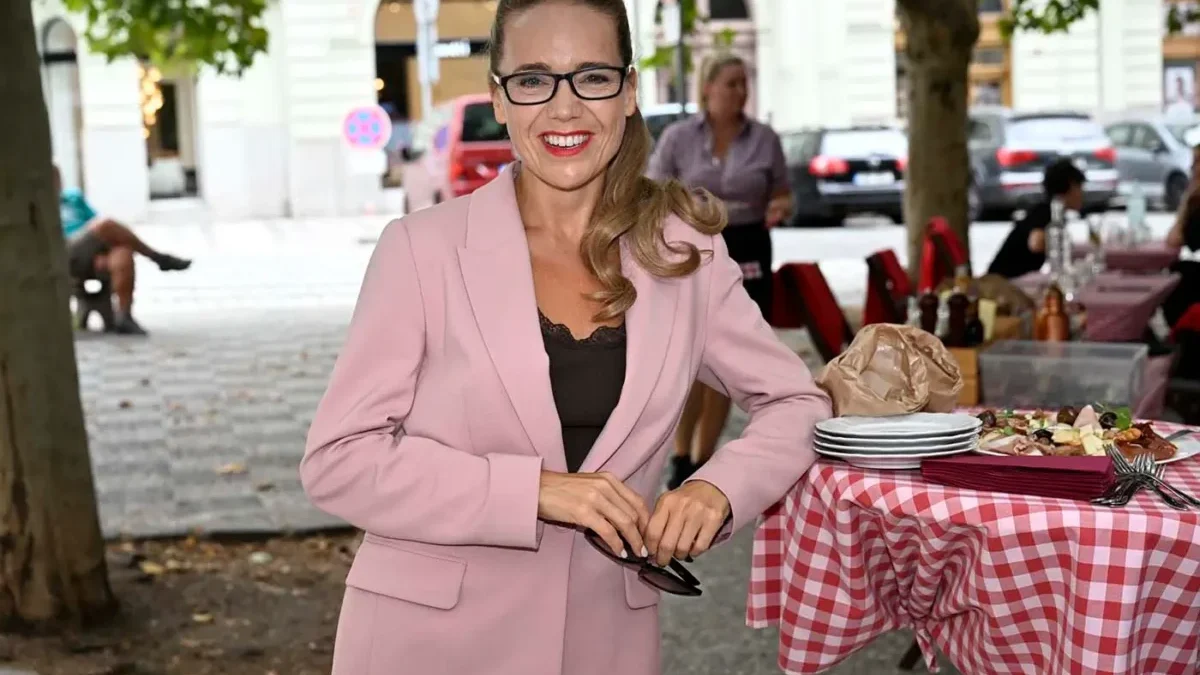 Žena s dlouhými vlasy, s brýlemi a v růžovém kostýmku stojí opřená o stůl s kostkovaným ubrusem