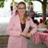 Žena s dlouhými vlasy, s brýlemi a v růžovém kostýmku stojí opřená o stůl s kostkovaným ubrusem