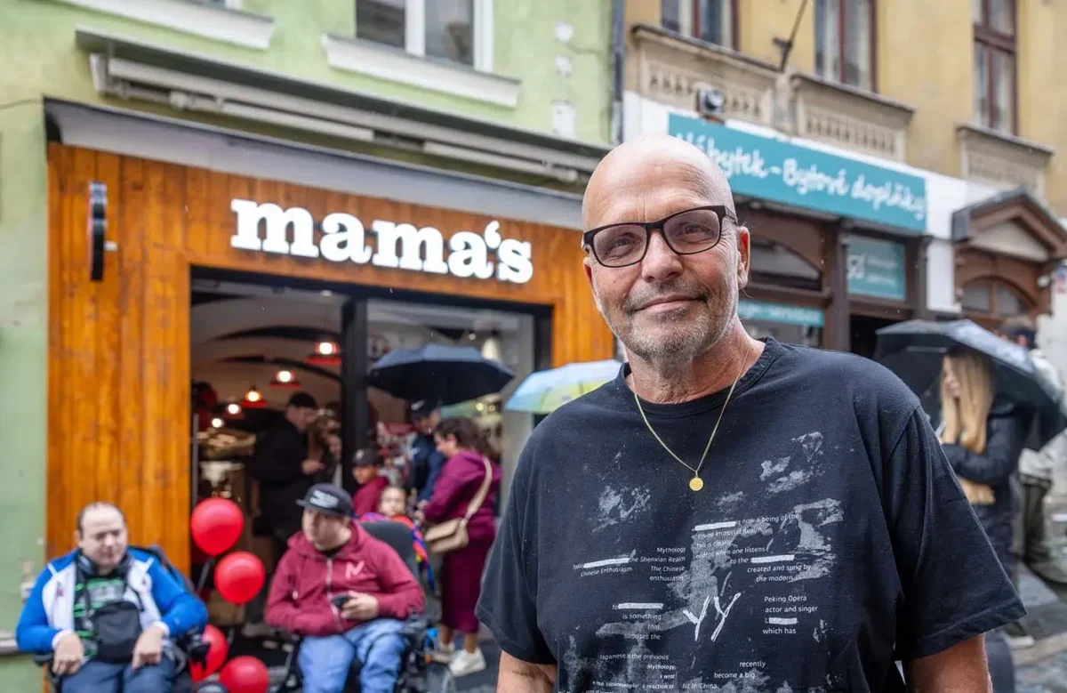 Starší muž bez vlasů a v tričku stojí venku na ulici a za ním jsou další lidé a podnik s bílým názvem nad vchodem