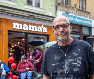 Starší muž bez vlasů a v tričku stojí venku na ulici a za ním jsou další lidé a podnik s bílým názvem nad vchodem